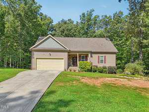 A single-story house with a sloped roof, two-car garage, and front porch. It's surrounded by a neat lawn and trees in the background. An American flag hangs near the porch.