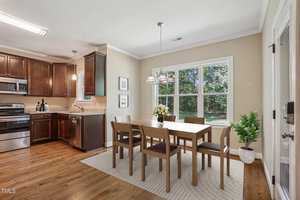 A kitchen and dining area with wooden flooring, featuring a dining table and six chairs on a rug. Stainless steel appliances and dark wood cabinets are in the kitchen. A window offers a view of trees, and a light fixture hangs over the table.