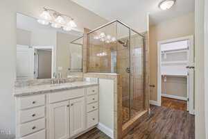Bright bathroom with a glass-enclosed shower, beige tiled walls, and wooden flooring. A white vanity with a granite countertop and two sink faucets is on the left. The door to a walk-in closet is visible in the background.