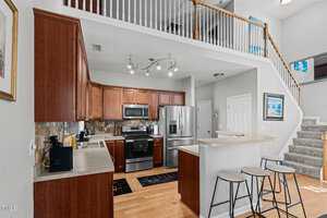 Modern kitchen with wooden cabinets, stainless steel appliances, and a breakfast bar with three stools. Overhead lighting illuminates the space. Stone backsplash and wooden floors add warmth. A staircase with white railing leads to an upper floor.