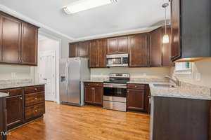A modern kitchen with wooden cabinets, stainless steel appliances, and granite countertops. The room features a hardwood floor and overhead lighting. A silver refrigerator, stove, and microwave are visible against the light-colored walls.