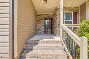 The image shows a house entrance with a covered porch area. The porch is accessed by a few brick steps and has a stone facade. The house number "2485" is displayed on a white column. Bushes are visible beside a window with red shutters.