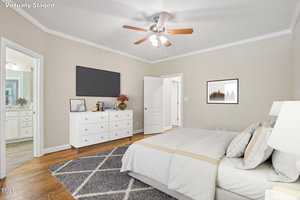 A virtually staged bedroom with a bed, dresser, and TV. The bed has patterned pillows and a beige throw blanket. A ceiling fan is above, and the room features hardwood floors. An open door leads to a bathroom with a visible vanity.