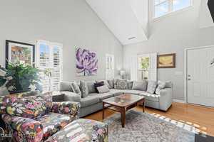 A bright living room with a large gray sectional sofa and a glass-topped coffee table. Two colorful floral armchairs face the sofa. Sunlight streams through plantation shutters, illuminating the hardwood floor and wall art, including a purple flower painting.