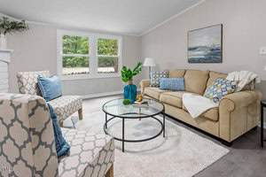 A cozy living room with a beige sofa, patterned armchairs, and colorful cushions. A glass coffee table sits on a white rug, holding decor items. A painting hangs on the wall, and large windows offer a view of greenery.