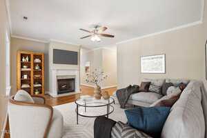 A cozy living room with a gray sectional sofa and navy blue pillows. A round glass coffee table sits on a light rug. The room has a fireplace, ceiling fan, bookshelf, and wall-mounted TV. Neutral walls and hardwood floors complement the decor.