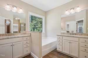 A bright bathroom with two vanity sinks, a large mirror, and overhead lights. A built-in bathtub is centered under a window with a view of green trees. The room features beige walls and wooden flooring.