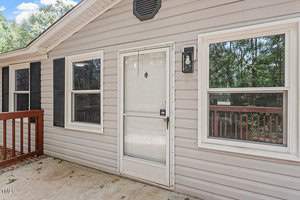 Single-story house exterior with beige siding, a glass front door, and two windows. A wooden porch railing is visible on the right. Surrounded by lush greenery under a clear sky.