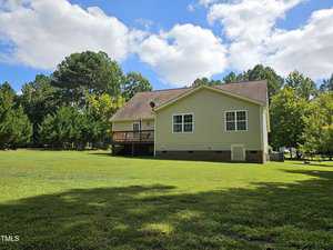 A beige house with a wooden deck sits in a spacious grassy yard surrounded by trees under a partly cloudy blue sky.