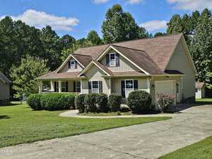 A suburban house with a beige exterior, gabled roof, and two dormer windows. A wide driveway leads up to the house, and neatly trimmed bushes line the front. The property is surrounded by lush green grass and tall trees in the background.