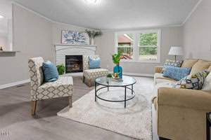 A cozy living room with light-colored walls and wood flooring. It features a beige sofa, patterned armchairs, and a circular glass coffee table. A fireplace is adorned with floral art and a potted plant. Large windows provide natural light.