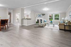 Spacious living room with light wood flooring, a beige sofa, patterned armchairs, and a round coffee table. A fireplace is on one wall, and large windows provide natural light. A dining area and kitchen are visible in the background.