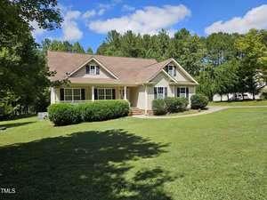 A charming one-story house at 2115 Turquoise Lane in Franklinton boasts a brown roof and beige siding on a spacious lawn. It features a front porch with white railings, enveloped by lush greenery and towering trees under a partly cloudy sky.