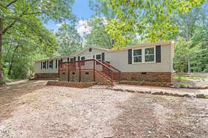 A single-story house with beige siding and a brick foundation is set among trees. It features multiple windows and a wooden deck with a railing. The property is surrounded by a mix of grass and bare earth, with a clear sky above.