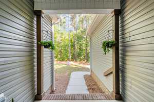 View from a covered entryway looking outside towards a tree-lined backyard. The pathway is flanked by two hanging planters with green foliage and bordered by light-colored siding walls.