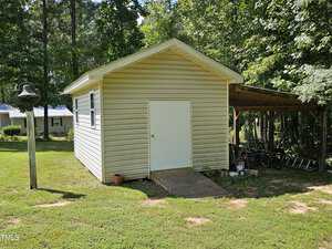 A small, light-colored shed with a gable roof sits on a grassy lawn. A rustic wooden structure is attached on the right, covering a couple of bicycles. A few trees are in the background, and a light fixture stands on the left.