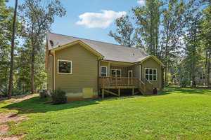A small, single-story house with a wooden deck sits on a grassy lawn surrounded by tall trees. The house has beige siding and a dark gray roof. The sky is clear and blue with a few clouds.