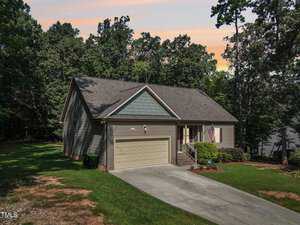 Nestled at 2485 Golden Forest Drive in Franklinton, this single-story house boasts a gray exterior, a two-car garage, and a charming flag by the entrance. Surrounded by trees and set against a sunset sky, it offers a concrete driveway leading right up to the welcoming garage.