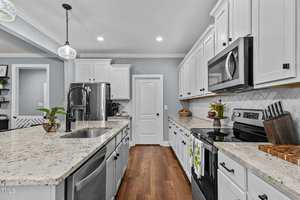 A modern kitchen with white cabinets, a stainless steel refrigerator, and a sleek black microwave. Granite countertops feature a sink, decorative plants, and utensils. Recessed lighting and pendant lights illuminate the space.