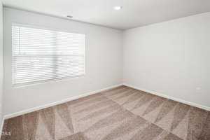 An empty room with white walls and beige carpet. A window with closed blinds is on the left, letting in soft natural light. The room has a simple ceiling light and visible outlet on the right wall. Carpet shows vacuum lines.
