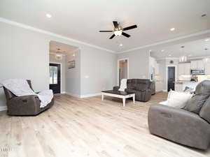 Spacious living room with light wood flooring, featuring two gray recliners, a matching sofa, and a white coffee table. Ceiling fan with lights overhead and an open view into a modern kitchen with white cabinetry and pendant lighting.