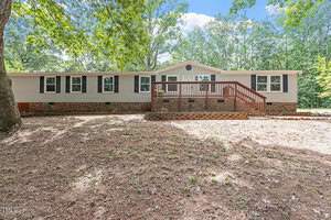 Located at 4032 Royal Oak Drive in Franklinton, this charming single-story house boasts light-colored siding and dark shutters. It features a wooden deck with stairs leading to the grassy yard, surrounded by large trees and a partly cloudy sky overhead.