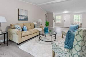 A cozy living room with a beige sofa, patterned armchairs, and decorative cushions. A round glass coffee table with a blue vase sits on a white rug. The walls are light gray with a seascape painting. Large windows and a white door are at the back.