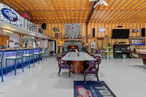 A spacious, rustic-themed bar area with a long marble table surrounded by chairs. The room features a wooden ceiling, a stone fireplace with a star above, several neon signs, a bar counter with stools, and vintage decor items.