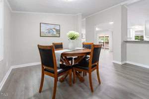 A modern dining room with a round wooden table and four black cushioned chairs. A white vase with flowers is on the table. Light grey walls and hardwood flooring. A landscape painting hangs on the wall, and there's an open view to the living area.