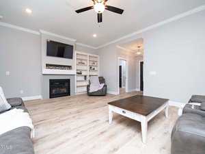 A modern living room with light wood floors, gray walls, and a ceiling fan. It features a fireplace with a wall-mounted TV, built-in shelves, leather armchairs, and a large coffee table in the center.