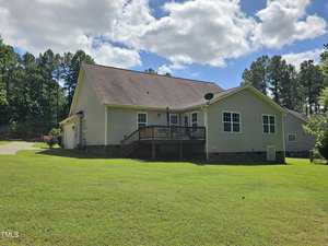 Rear view of a single-story house with tan siding, a wooden deck, and a sloped roof under a partly cloudy sky. The property includes a well-maintained grassy lawn bordered by trees.