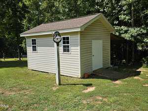 A small, white shed with a gray roof stands on a grassy area, surrounded by trees. A wooden ramp leads to a white door. In the foreground, there's a bell mounted on a wooden post.