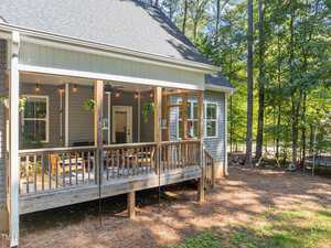 A wooden patio extends from a house, featuring a screened-in porch with hanging plants and outdoor seating. Surrounded by tall trees and located in a sunny, wooded area, the scene conveys a tranquil, natural setting.