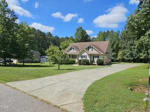 A charming house with beige siding and a sloped roof sits at the end of a long driveway, surrounded by lush green lawns and tall trees. The sky above is clear and blue with a few scattered clouds.