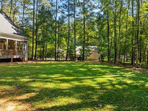 A sunny backyard features a spacious lawn surrounded by tall trees. A house with a covered porch is on the left, and another house is partially visible in the background, framed by the lush greenery of the wooded area.