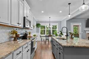 A modern kitchen with white cabinets, light granite countertops, and stainless steel appliances. A large island with a sink and a drop-leaf table with chairs are present. Green plants decorate the space. Large windows provide natural light.