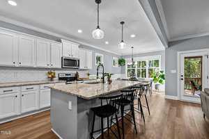 A modern kitchen with white cabinets and a large granite island with barstools. Pendant lights hang above the island. The room features a herringbone backsplash, stainless steel appliances, and large windows with greenery outside.
