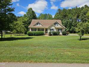 A charming, single-story house with a gable roof sits amid a spacious green lawn under a partly cloudy blue sky. Trees and shrubs border the property, adding to the serene, suburban setting.