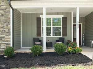 A cozy front porch with two woven chairs and a small table. The house has green siding, white columns, and a vertical "Welcome" sign. Potted flowers and shrubs decorate the area, and the number "3607" is displayed on a column.