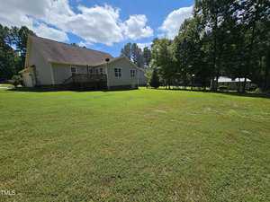 A light gray house with a wooden deck is surrounded by a large, green grassy yard. Tall trees border the property under a blue sky with scattered clouds.