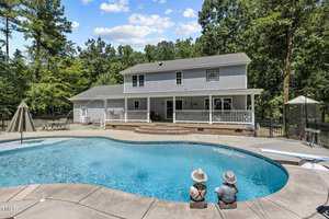 A spacious two-story house with a large backyard featuring a swimming pool. Two decorative figures resembling children sit at the pool's edge. The area is surrounded by lush trees and a patio with furniture is visible under a covered porch.
