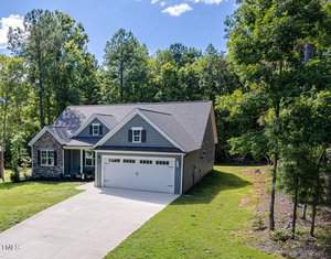 A single-story house with a large garage sits in a wooded area. The home has a gabled roof, stone accents, and a concrete driveway. Surrounded by lush green trees under a clear blue sky, it exudes a serene and natural setting.