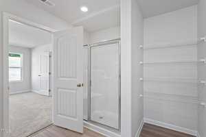 A bright, empty bathroom with a glass shower door, adjacent to a walk-in closet with wire shelving. The space has white walls, a wood floor, and a doorway leading to a carpeted room with a window.