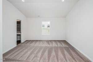 An empty room with white walls and a beige carpet featuring geometric vacuum lines. There's a single window in the far wall, allowing natural light. An open door on the left leads to another room with visible bathroom fixtures.
