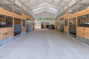 Interior of a modern horse stable with metal and wood construction. The stable features multiple stalls on both sides of a wide, concrete center aisle. Bright natural light comes in through the open doors at the far end.