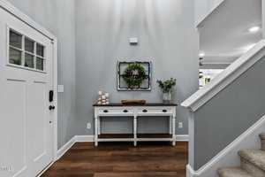 A hallway with a white front door and a wooden console table. The table features decorative items including a basket of books, a candle holder, and a plant. A wreath is centered above the table, and a staircase is visible to the right.