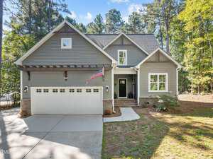 Nestled at 3714 Sapphire Court in Franklinton, this gray house boasts a large driveway and attached garage. The welcoming porch features a wreath on the door, while an American flag proudly hangs above the entrance. Tall trees surround the home under a clear blue sky.