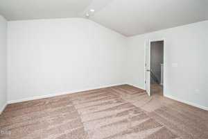 A vacant room with clean beige carpeting and white walls. An open door leads to another room, revealing part of a staircase. The space is well-lit with natural light, and the ceiling is smooth and slightly vaulted.