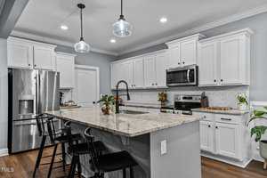 A modern kitchen with white cabinets and stainless steel appliances, including a refrigerator and oven. The large granite island features a sink and black chairs. Pendant lights hang from the ceiling, and small plants decorate the space.