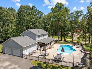 Aerial view of a two-story gray house with a spacious patio, outdoor seating, and a swimming pool. The property is surrounded by a black metal fence and lush trees, with a clear blue sky above.